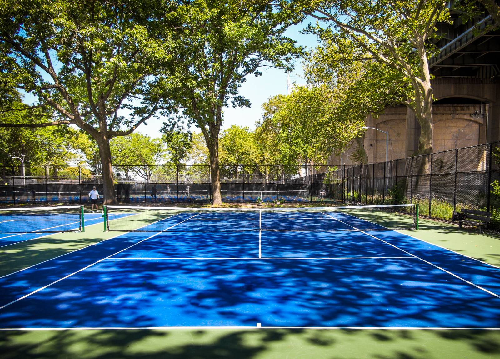 Astoria Park's public tennis courts shaded by mature trees, with the RFK Bridge piers visible at right. Players in the background.